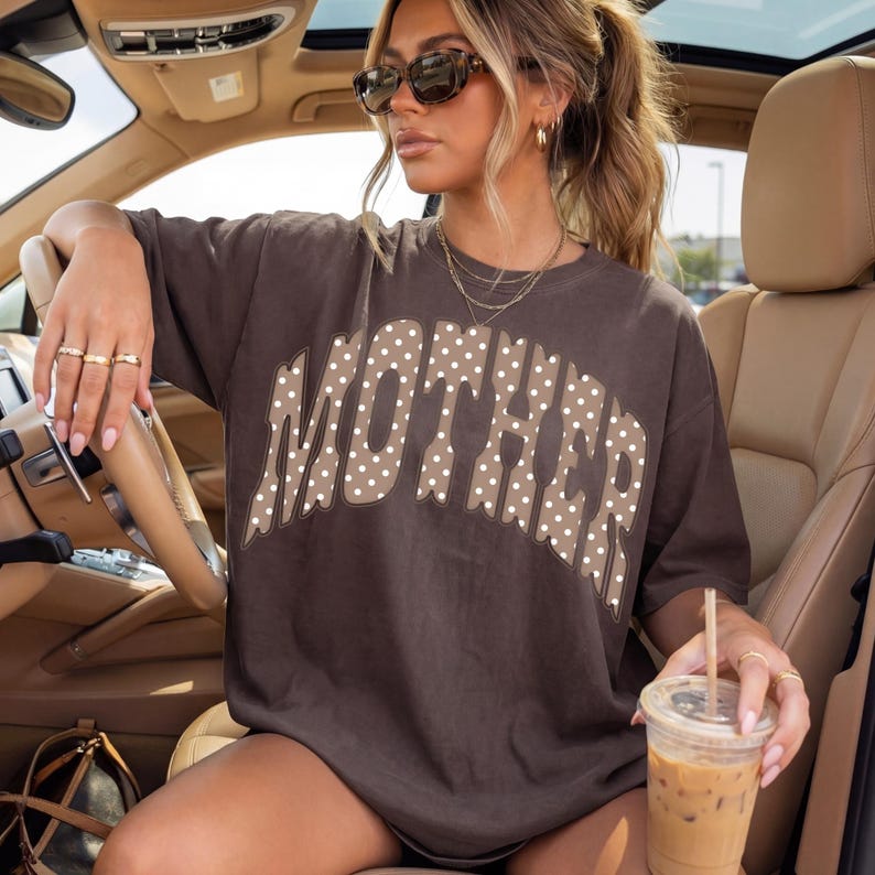 May include: Brown t-shirt with the word "MOTHER" in a dotted pattern. The shirt is oversized and has short sleeves. The model is wearing sunglasses and holding a beverage.