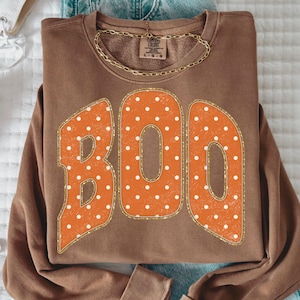 Könnte beinhalten: Ein braunes Langarm-Sweatshirt mit dem Wort BOO in großen orangefarbenen Buchstaben mit weißen Punkten und einer goldglitzernden Umrandung. Das Kleidungsstück hat einen Rundhalsausschnitt und ein Comfort Colors Markenetikett am Innenkragen.