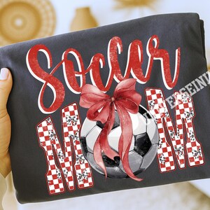 May include: Dark gray t-shirt with the words "Soccer Mom" in red script and checkered letters. A soccer ball with a red bow is in the center. The shirt is folded and held by a hand.