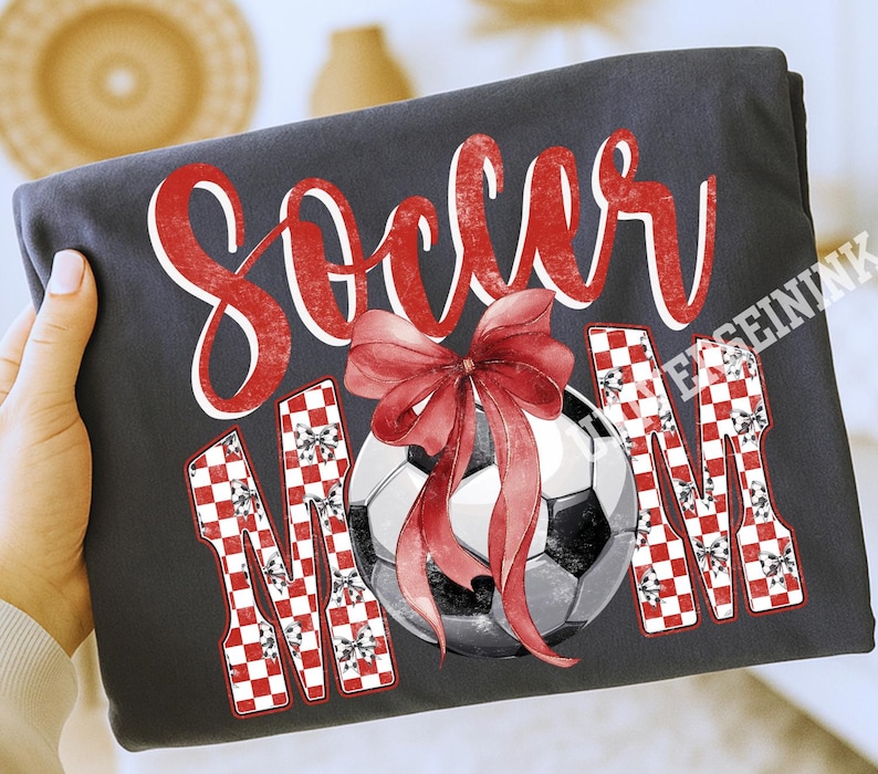 May include: Dark gray t-shirt with the words "Soccer Mom" in red script and checkered letters. A soccer ball with a red bow is in the center. The shirt is folded and held by a hand.