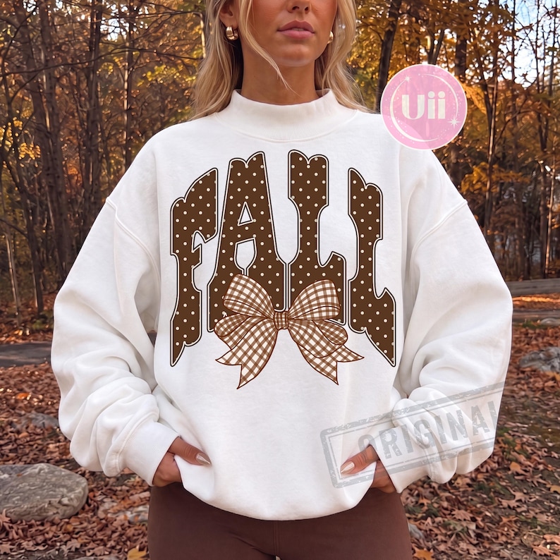 May include: A white long-sleeve sweatshirt featuring a mock neck and a large graphic on the front. The graphic displays the word FALL in brown letters with white polka dots, accented by a brown and white gingham patterned bow at the center.