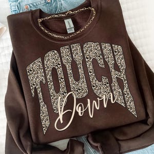 May include: A dark brown crewneck sweatshirt featuring the text TOUCH DOWN. The word TOUCH is printed in a large, blocky font with a leopard print pattern, while the word DOWN is written below in a white, cursive script font.