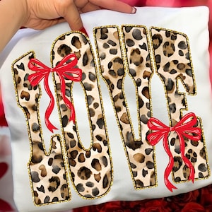 Könnte beinhalten: Weißes Kissen mit dem Wort "LOVE" in Leopardenmuster, umrandet mit goldenem Glitzer. Rote Schleifen schmücken die Buchstaben. Das Kissen wird von einer Person gehalten.