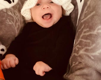 May include: A baby wearing a white crocheted ghost hat with black eyes and a black long-sleeved shirt. The baby is lying on a gray and white blanket with a ghost pattern.