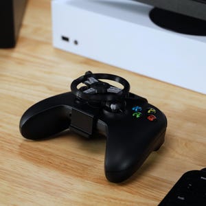 May include: A black Xbox controller with a black and gray steering wheel attachment. The controller is on a wooden surface.