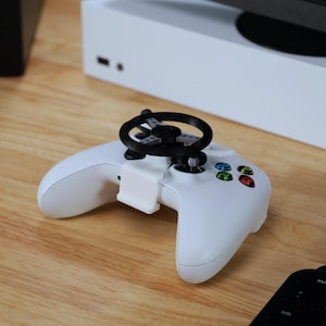 May include: A white Xbox controller with a black steering wheel attachment. The controller is on a wooden surface.