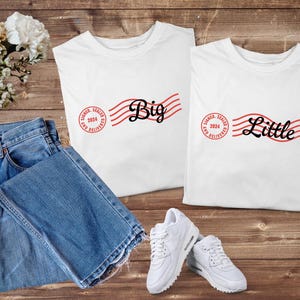 May include: Two white t-shirts with the words "Big" and "Little" in a script font, accompanied by red wavy lines and a circular stamp that reads "Signed, Sealed, and Delivered 2024." Blue jeans and white sneakers complete the casual ensemble.