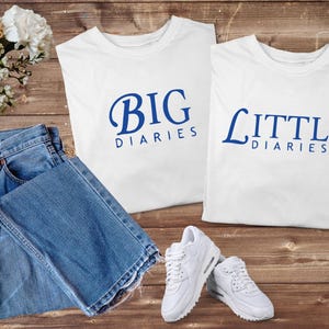 May include: Two white t-shirts with the text "BIG DIARIES" and "LITTLE DIARIES" in blue script. A pair of blue jeans and white sneakers are also in the image. The items are arranged on a wooden surface.
