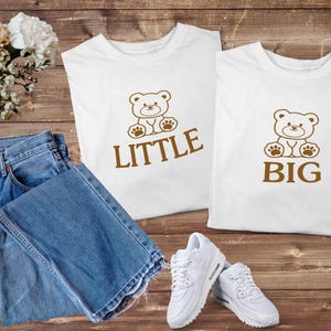 Puede incluir: Dos camisetas blancas con gráficos de osos de peluche marrones y las palabras "LITTLE" y "BIG". Un par de vaqueros azules y zapatillas blancas también están en la imagen. Los artículos están colocados sobre una superficie de madera.