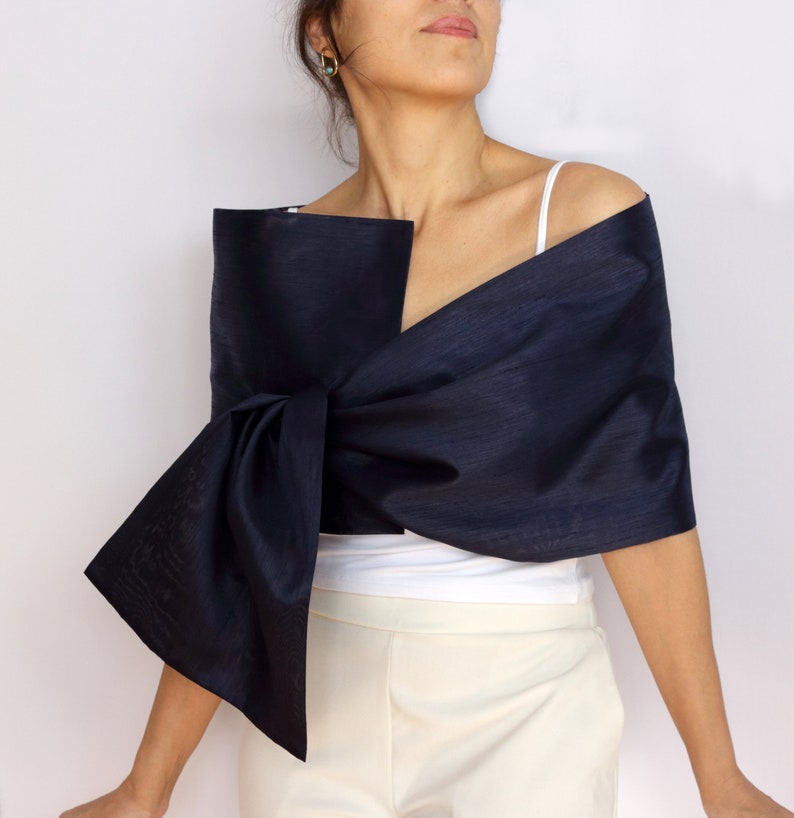 Pu&ograve; includere: Uno scialle blu navy con un grande dettaglio a fiocco. Lo scialle &egrave; drappeggiato sulle spalle. La persona indossa un top bianco e pantaloni color crema. Il tessuto &egrave; lucido.