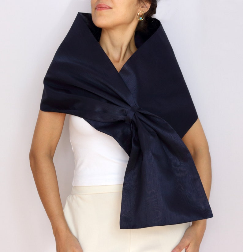 Pu&ograve; includere: Uno scialle blu navy con un grande dettaglio a fiocco. Lo scialle &egrave; realizzato in un tessuto lucido ed &egrave; drappeggiato sulle spalle. La modella indossa un top bianco e pantaloni color crema.