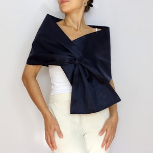 Pu&ograve; includere: Uno scialle blu navy con un design a forma di fiocco. Lo scialle &egrave; drappeggiato sulle spalle e sul petto. Il tessuto ha una lucentezza sottile. La modella indossa un top bianco e pantaloni color crema.