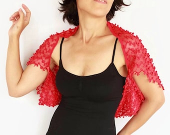 May include: A bright red lace bolero shrug featuring a delicate floral pattern with small raised textured details. The garment has short sleeves and an open front design, crafted from a sheer, lightweight fabric that drapes over the shoulders.