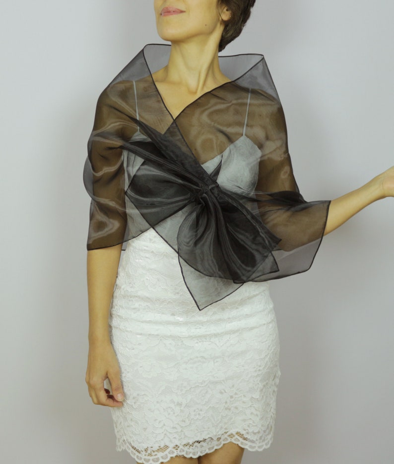 May include: A black sheer shawl with a large bow in the front. The shawl is draped over a white lace dress.