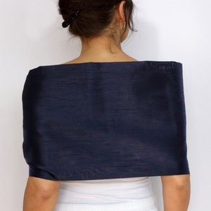 Pu&ograve; includere: Uno scialle di seta blu navy drappeggiato sulle spalle. Lo scialle ha un aspetto liscio e strutturato e una forma rettangolare. La persona indossa un top bianco sotto.