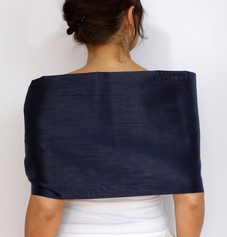 Pu&ograve; includere: Uno scialle di seta blu navy drappeggiato sulle spalle. Lo scialle ha un aspetto liscio e strutturato e una forma rettangolare. La persona indossa un top bianco sotto.