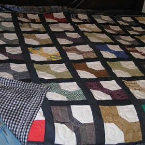 May include: A quilt made of various fabric scraps in a patchwork design. The quilt is made up of squares with a bow tie shape in the center of each square. The squares are sewn together with black fabric. The quilt is on a bed with a blue and white checkered sheet.