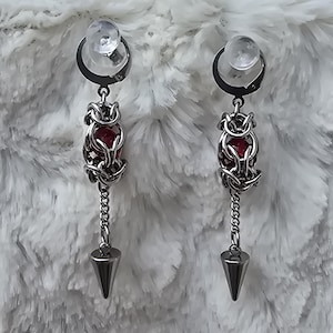 May include: A pair of silver-toned earrings with a clear, round stud at the top. The earrings feature a woven design around a red gem, with a chain and a silver cone at the bottom. The background is a fluffy, white material.