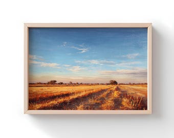 Op de afbeelding: Een ingelijst landschapsschilderij toont een veld met gouden tarwe onder een blauwe lucht met verspreide wolken. Een zandweg leidt in de verte, met bomen aan de horizon. Het kunstwerk zit in een lichtgekleurde houten lijst.