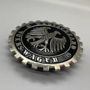 Puede incluir: Un emblema metálico plateado con un centro negro que presenta un diseño de águila. El emblema es circular con un borde en forma de engranaje y las palabras "AUTOS DEUTSCH CLUB" y "WAGEN" están inscritas.