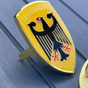 May include: A gold-colored metal car emblem featuring the German coat of arms. The emblem has a yellow shield with a black eagle and red crosses. The text "DEUTSCHLAND" is written in black letters around the top of the shield.