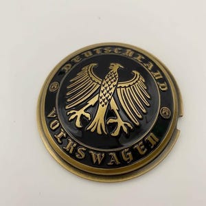 Op de afbeelding: Een rond, bronskleurig embleem met de woorden "VOLKSWAGEN" en "DEUTSCHLAND" rond een goudkleurige adelaar op een zwarte achtergrond. Het embleem heeft een licht verhoogd ontwerp.