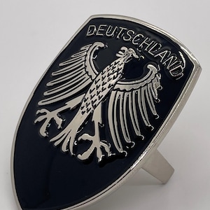 Peut inclure: Bague noire et argentée représentant les armoiries allemandes, un aigle et le mot "DEUTSCHLAND" en lettres argentées. La bague a une face en forme de bouclier avec une finition polie.