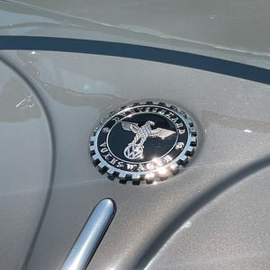 Może przedstawiać: Zbliżenie srebrnego emblematu Volkswagena z szczegółowym wzorem orła i napisem "VOLKS WAGEN" w okrągłym wzorze. Emblemat umieszczony jest na szarej powierzchni samochodu, odbijając światło słoneczne.