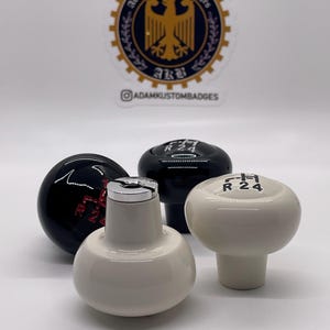 Puede incluir: Tres pomos de palanca de cambios, dos negros y uno blanco. El pomo blanco tiene letras negras que dicen "R 24".