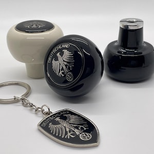 Op de afbeelding: Drie zwarte en witte versnellingspookknoppen met een zilveren adelaar en het woord "DEUTSCHLAND" erop. Eén knop is wit met een zwarte adelaar en de andere twee zijn zwart met een zilveren adelaar. Er is ook een zwarte en zilveren sleutelhanger met dezelfde adelaar en tekst.