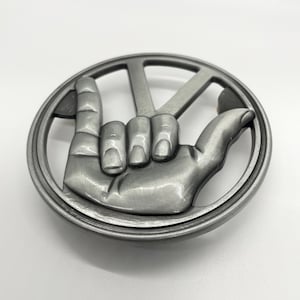 Może przedstawiać: Srebrna metalowa klamra do paska przedstawiająca gest dłoni z wyciągniętym palcem wskazującym i małym palcem, tworzącym symbol 'rock'. Dłoń znajduje się w okrągłej ramie z kształtem 'V' nad palcami.
