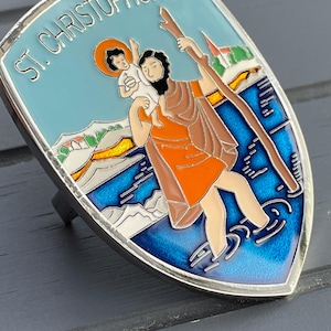 Op de afbeelding: Een zilveren, schildvormige speld met een blauw, wit en oranje emaille ontwerp. De speld toont een afbeelding van Sint Christoffel die een kind over een rivier draagt. De tekst "ST. CHRISTOPHORUS" is in wit op een blauwe achtergrond geschreven.