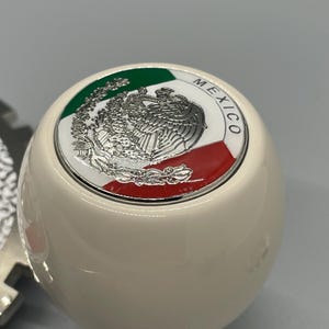 May include: Cream-coloured shift knob with a metallic emblem featuring the Mexican coat of arms. The emblem is bordered by green, white, and red, the colours of the Mexican flag. The knob has a smooth, rounded shape.