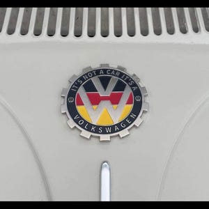 May include: Close-up of a classic Volkswagen emblem. The emblem is circular with the words "VOLKSWAGEN" and "IT'S NOT A CAR IT'S A" in a silver border. The VW logo is in the centre, with red, black, and yellow colours.