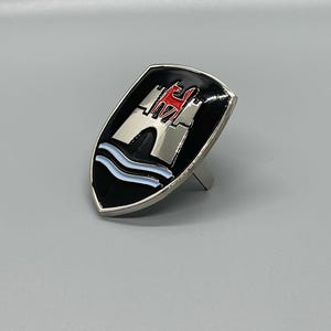 Puede incluir: Un emblema de metal plateado y negro con un castillo y un techo rojo, una ola azul y un fondo negro.