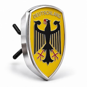 Może przedstawiać: Metalowy emblemat w kształcie tarczy z żółtym tłem, czarnym orłem pośrodku i napisem DEUTSCHLAND srebrnymi literami u góry. Emblemat posiada srebrną obwódkę oraz dwa czarne bolce wystające z tylnej części przedmiotu.