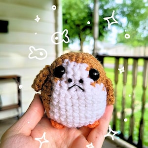 May include: A handmade crochet Porg plushie. The small, round creature has a brown head, white body, black eyes, and orange feet. The plushie is held in a hand, with a blurred green background.