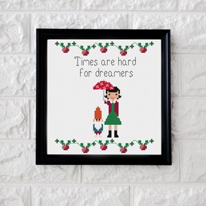 May include: Cross-stitch art in a black frame. The artwork features a woman holding a red umbrella with a gnome. The text "Times are hard for dreamers" is above the figures. Green and red floral accents border the top and bottom.