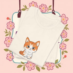 May include: Two cream-coloured t-shirts are stacked, each featuring a cartoon cat illustration in orange and white. The shirts are surrounded by a floral design with pink flowers and green leaves, set against a pink background.