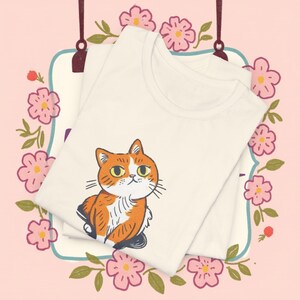 May include: Two cream-coloured t-shirts are stacked, each featuring a cartoon cat illustration in orange, white, and black. The shirts are set against a pink background with floral and leaf accents, creating a whimsical and playful design.
