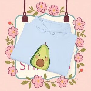 Pode incluir: Um moletom azul claro com um desenho de abacate de desenho animado. O abacate tem uma expressão de surpresa e é verde com um caroço marrom. O moletom é dobrado, em um fundo rosa com uma borda floral.