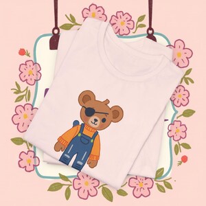 May include: White t-shirt featuring a cartoon bear wearing an eye patch, orange sweater, and blue overalls. The bear has a backpack. The shirt is folded on a pink background with floral accents.