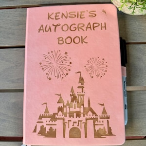 K&ouml;nnte beinhalten: Ein hellrosa Autogrammbuch mit dem eingravierten Text KENSIE'S AUTOGRAPH BOOK, einer gro&szlig;en Burgsilhouette und zwei Feuerwerksmotiven. Ein schwarzer Permanentmarker steckt an der Seite des Buches, das zwei d&uuml;nne Lesezeichenb&auml;nder enth&auml;lt.