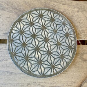 May include: Round wooden coaster with a geometric starburst pattern. The coaster is light gray with a darker gray border and etched design. The coaster is approximately 4 inches in diameter.
