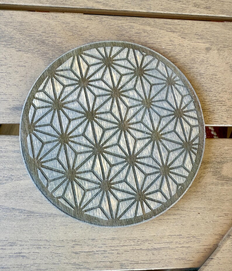 May include: Round wooden coaster with a geometric starburst pattern. The coaster is light gray with a darker gray border and etched design. The coaster is approximately 4 inches in diameter.