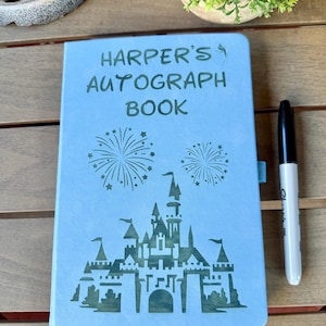 K&ouml;nnte beinhalten: Ein hellblaues Autogrammbuch mit dem dunkelgr&uuml;nen Text "HARPER'S AUTOGRAPH BOOK" &uuml;ber einer Grafik eines Schlosses und zwei Feuerwerken. Ein schwarzer Stift liegt rechts neben dem Buch, das ein blaues Lesezeichenband besitzt.