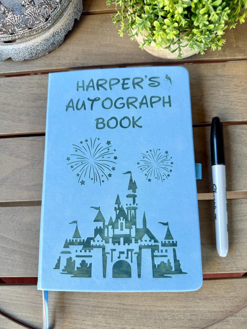 K&ouml;nnte beinhalten: Ein hellblaues Autogrammbuch mit dem dunkelgr&uuml;nen Text "HARPER'S AUTOGRAPH BOOK" &uuml;ber einer Grafik eines Schlosses und zwei Feuerwerken. Ein schwarzer Stift liegt rechts neben dem Buch, das ein blaues Lesezeichenband besitzt.