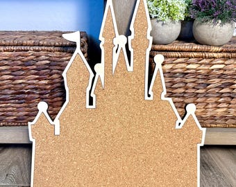 May include: A corkboard in the shape of a castle with a white border and flag details. The castle design features multiple turrets and a rectangular corkboard base. The background includes decorative plants and a woven basket.