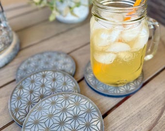 May include: A refreshing drink in a mason jar with ice and a yellow straw sits on a gray geometric patterned coaster. Three additional coasters are visible, along with a small potted plant. The coasters have a blue trim and are on a wooden table.
