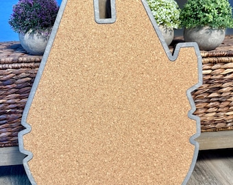 May include: A cork coaster in the shape of the Millennium Falcon spaceship. The coaster has a light brown cork center with a gray border. The coaster is sitting on a wooden surface.